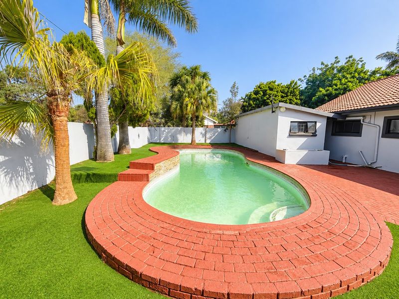 Outdoor pool area featuring a curved brick surround, artificial grass landscaping, and perimeter walling, set against a residential building with a tiled roof.