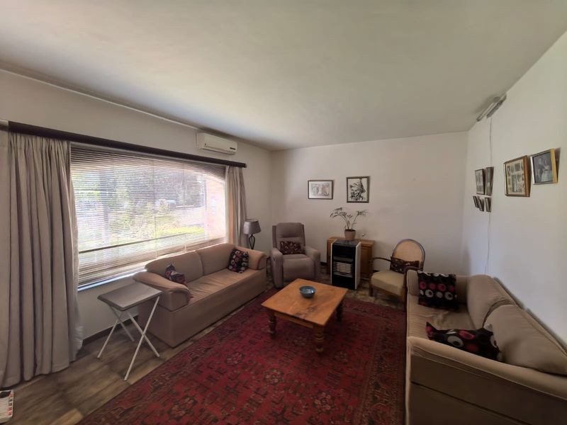 A comfortable living room featuring laminate flooring, a wall-mounted air conditioning unit, and large windows with venetian blinds and curtains.