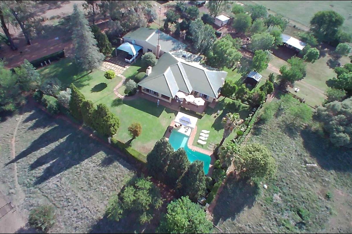 Aerial view of a farm property featuring a main residence with a metal roof, swimming pool, paved patio area, and surrounding landscaped grounds.