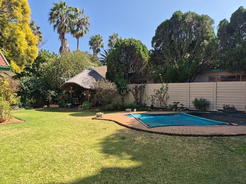A private backyard featuring a swimming pool with a cover, a traditional thatched lapa, and a well-maintained lawn area.