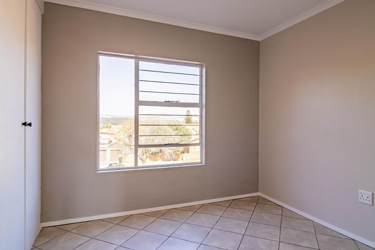 A bright, empty bedroom featuring neutral wall tones, tiled flooring, and a built-in cupboard with secure window burglar bars.
