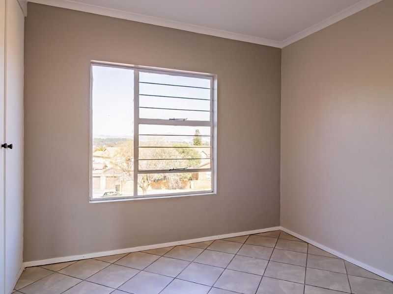A bright, empty bedroom featuring neutral wall tones, tiled flooring, and a built-in cupboard with secure window burglar bars.