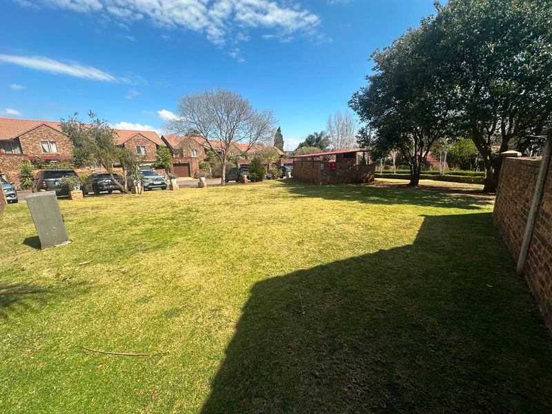 A spacious communal garden area featuring a well-maintained lawn, mature trees, and brick boundary walls within a townhouse complex.