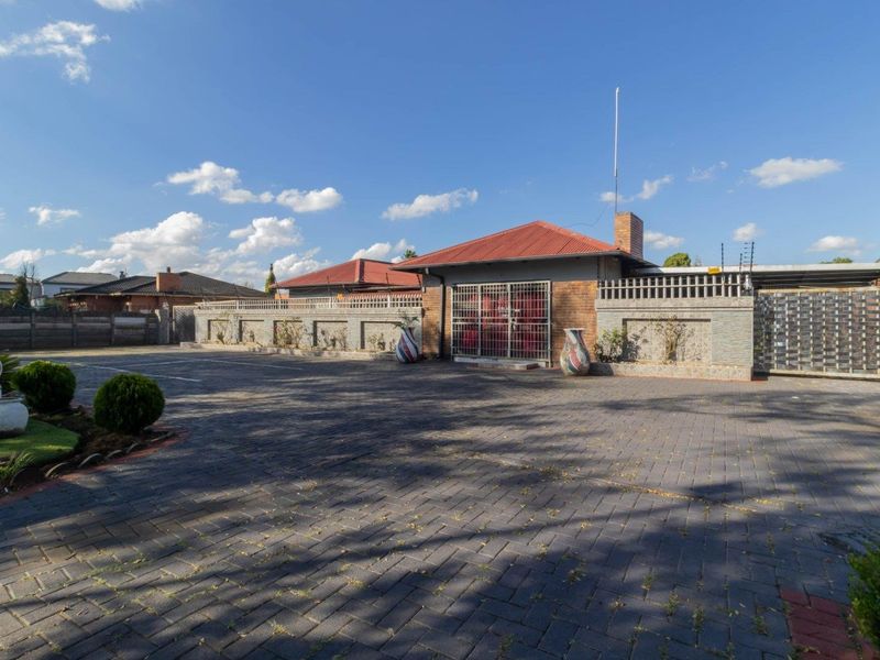 Front exterior view of the property featuring a large paved driveway, secure boundary wall with electric fencing, and a metal roof structure.