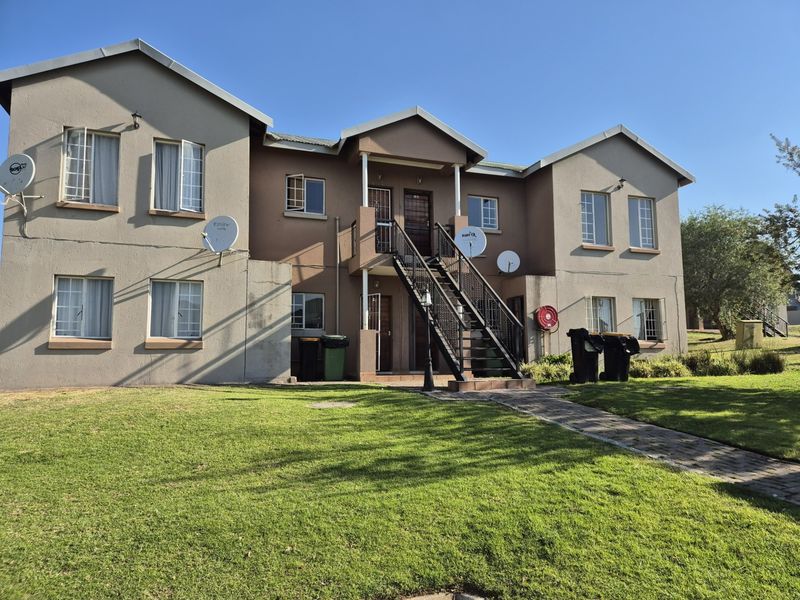 Rear view of a multi-story apartment complex featuring a central metal staircase, paved access path, and established lawn area.