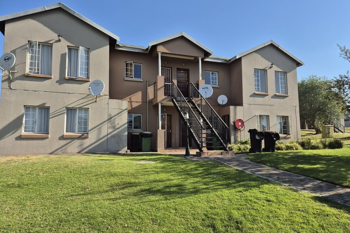 Rear view of a multi-story apartment complex featuring a central metal staircase, paved access path, and established lawn area.