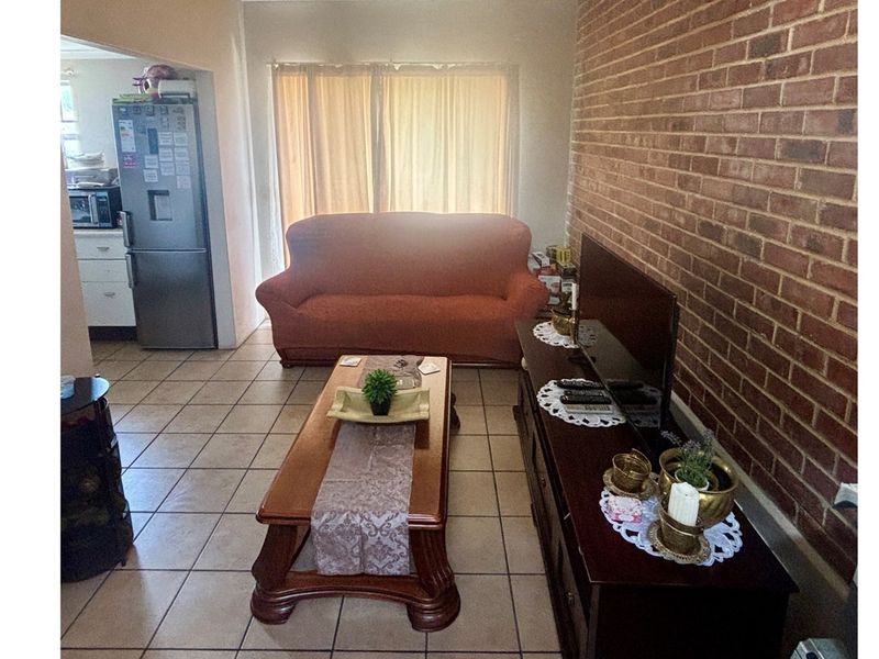 A living room featuring an exposed brick feature wall and ceramic floor tiles, with an open-plan layout connecting to the kitchen area.