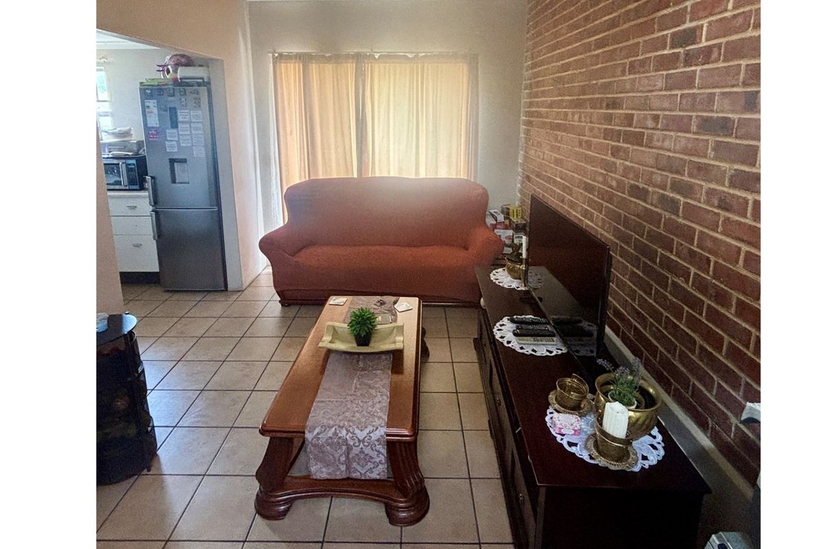 A living room featuring an exposed brick feature wall and ceramic floor tiles, with an open-plan layout connecting to the kitchen area.