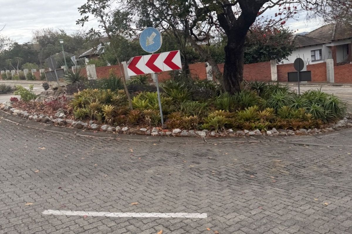 A view of a paved roundabout featuring a landscaped traffic island with a mature tree and brick boundary wall in the background.