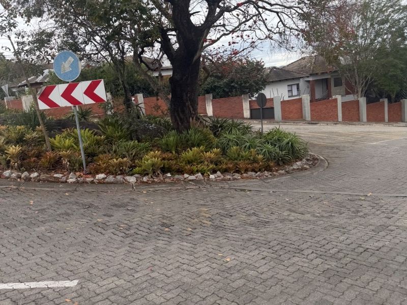 A view of the paved street approach featuring a landscaped traffic island with a mature tree and brick boundary wall.