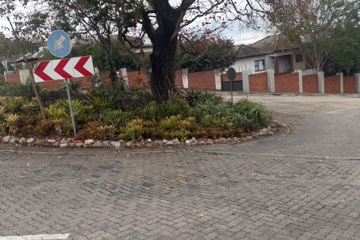 A view of the paved street approach featuring a landscaped traffic island with a mature tree and brick boundary wall.