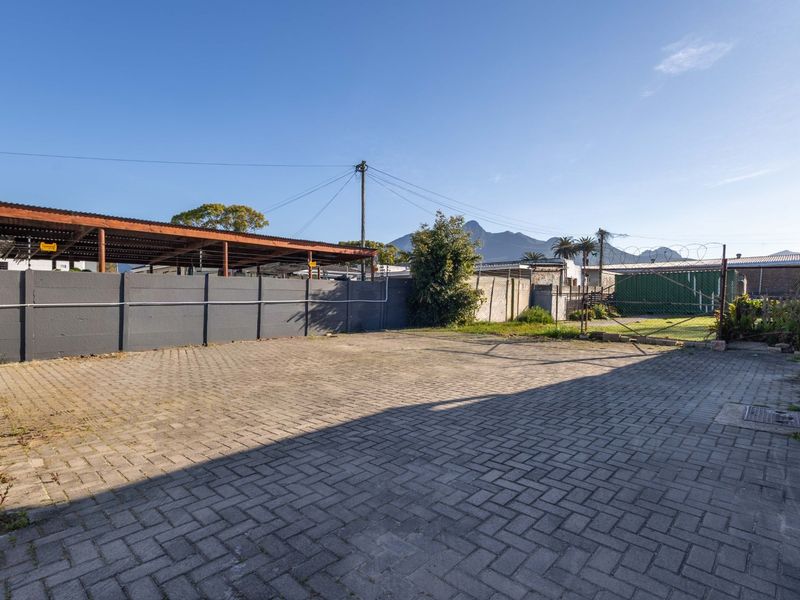 A spacious paved rear yard area featuring a perimeter wall with security fencing, a metal gate, and a covered parking structure.