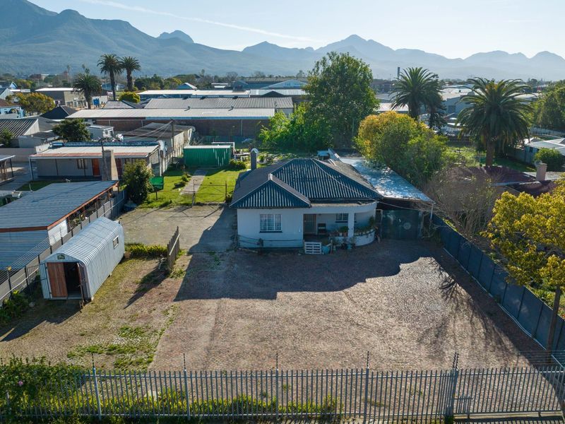 Aerial view of a commercial property featuring a main building, large paved driveway, metal perimeter fencing, and a detached corrugated metal storage shed.