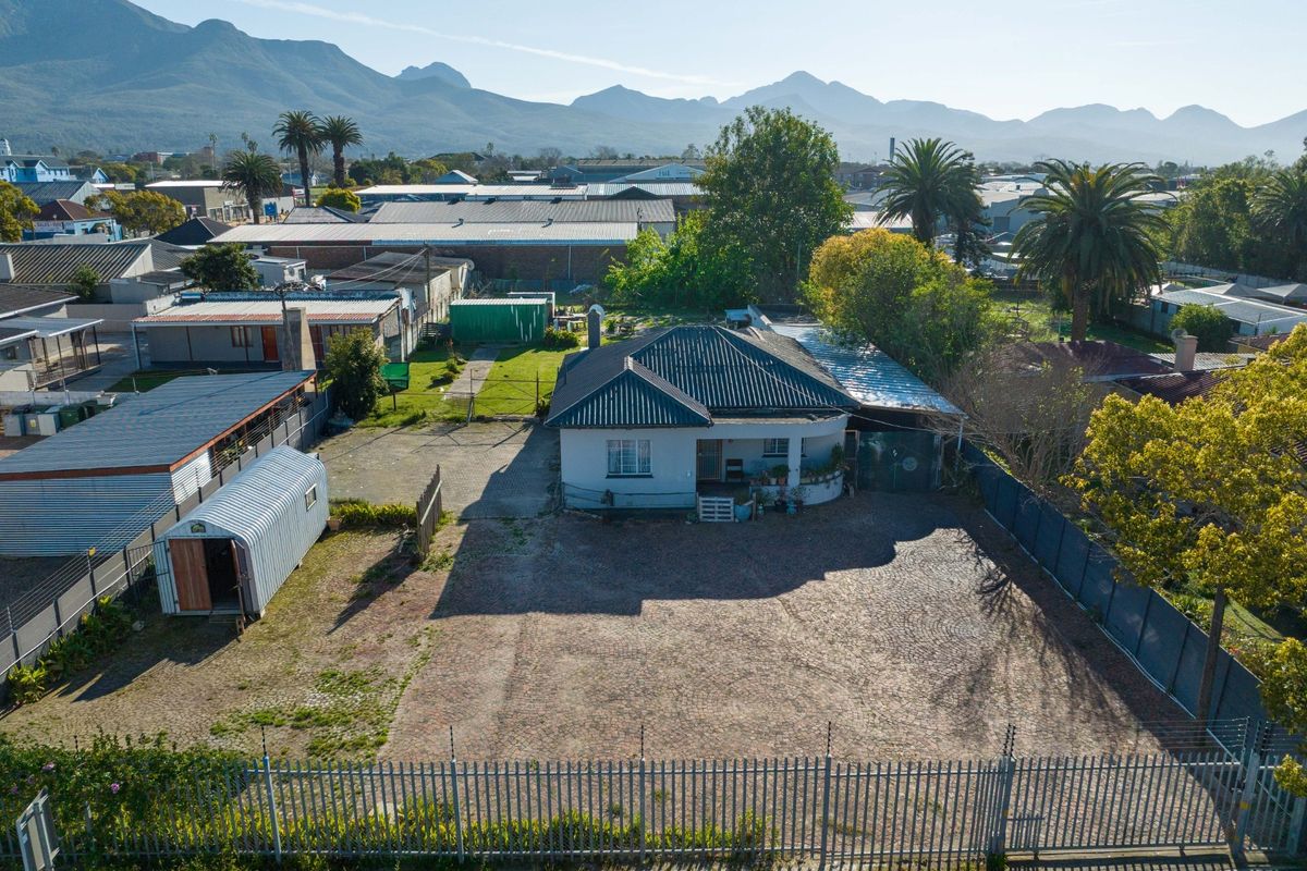 Aerial view of a commercial property featuring a main building, large paved driveway, metal perimeter fencing, and a detached corrugated metal storage shed.