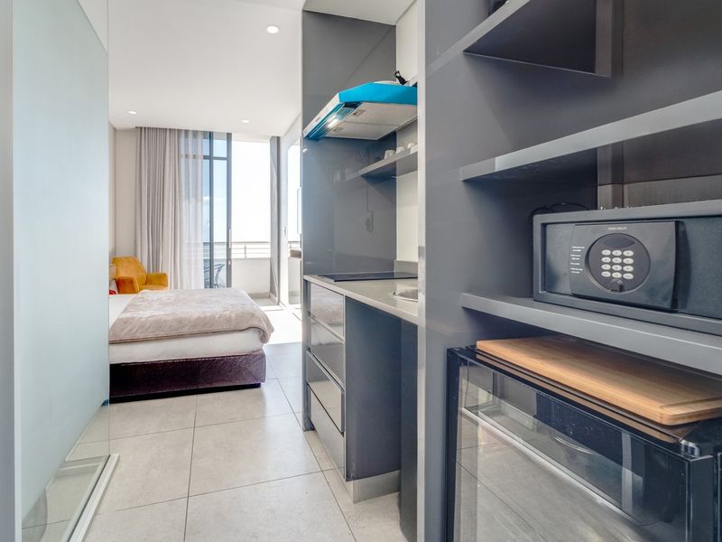 Modern open-plan studio apartment featuring a compact kitchenette with built-in cabinetry, hob, and extractor fan, leading to a bedroom area with balcony access.