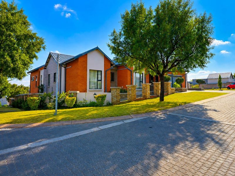 A well-maintained single-story residential house featuring a brick facade, metal roofing, and stone pillar fencing, situated on a paved street with landscaped grounds.
