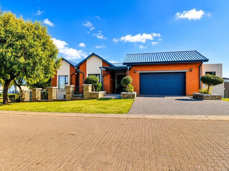 A well-maintained single-story brick house featuring a metal roof, double garage, paved driveway, and landscaped front garden with stone masonry accents.