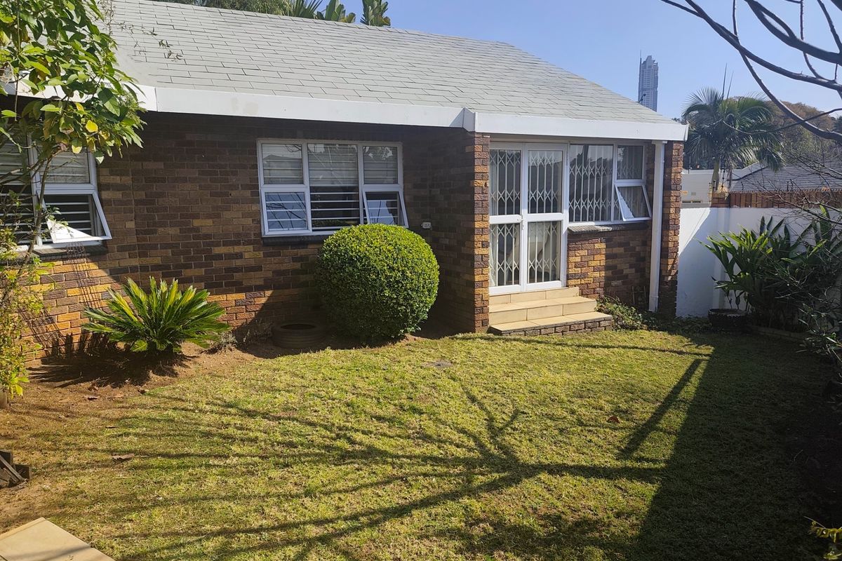 A rear view of a brick-faced house featuring a small lawn, security gates, and aluminium window frames.