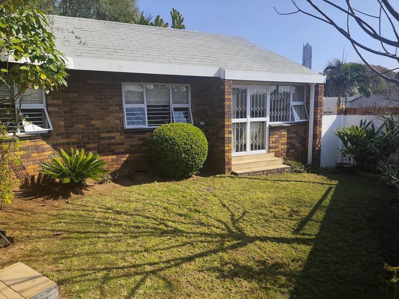 A rear view of a brick-faced house featuring a small lawn, security gates, and aluminium window frames.