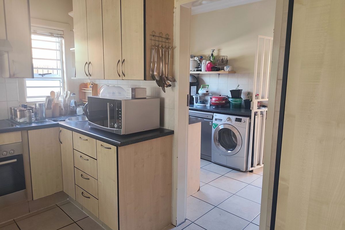 A functional kitchen featuring light wood-finish cabinetry, dark laminate countertops, and tiled flooring, with an adjacent scullery area visible through the doorway.