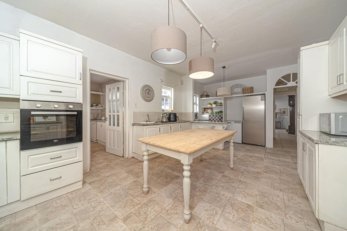 A spacious kitchen featuring white shaker-style cabinetry, stone countertops, tiled flooring, and integrated oven, with an adjacent scullery area visible through the doorway.