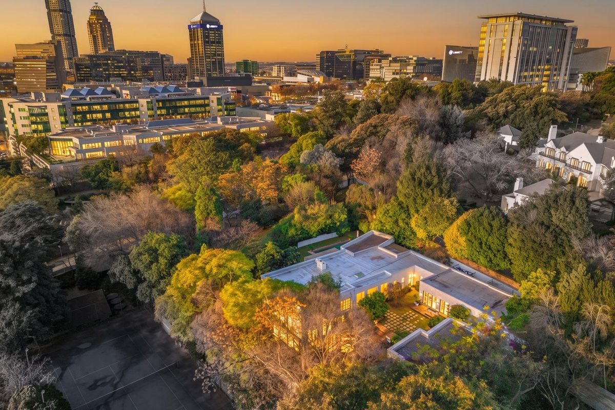 Aerial view of a commercial property featuring a tennis court, landscaped gardens, and a flat-roofed building set against a city skyline backdrop.