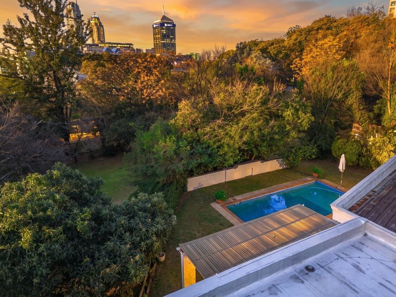 Aerial view of a property featuring a swimming pool, landscaped garden, and boundary wall, set against a scenic urban skyline backdrop.