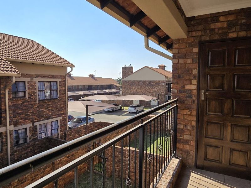 A covered balcony featuring exposed brick walls, a wrought iron balustrade, and a timber entrance door, overlooking the apartment complex grounds.