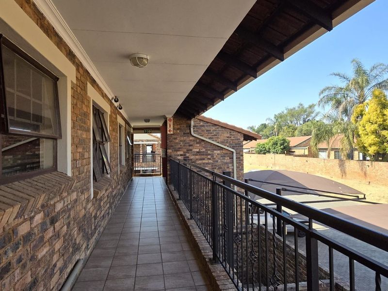 An exterior covered walkway featuring exposed brick walls, tiled flooring, and a metal balustrade overlooking the apartment complex grounds.