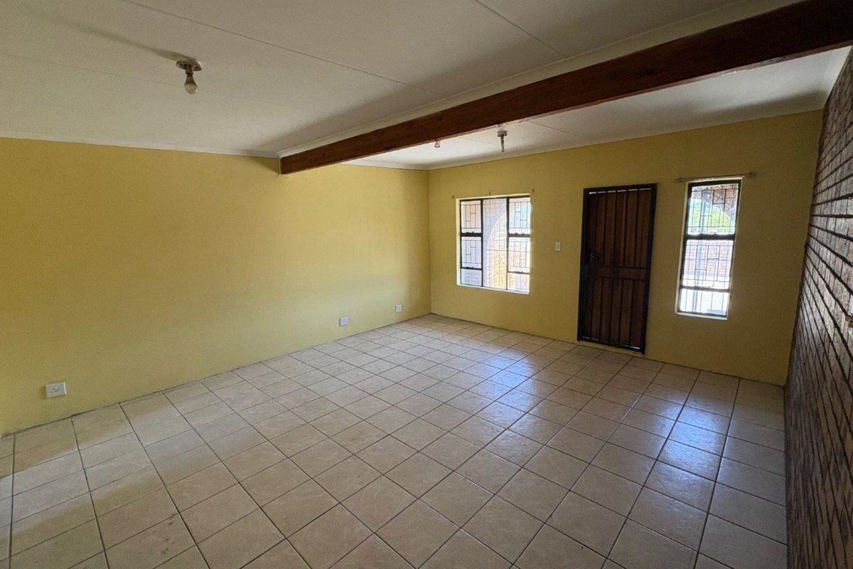 Spacious, empty living area featuring tiled flooring, a decorative exposed timber ceiling beam, and a textured brick feature wall with secure window access.