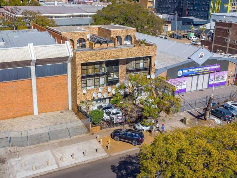 Aerial view of a multi-story commercial brick building featuring arched windows, flat roof, and secure perimeter fencing in an urban setting.