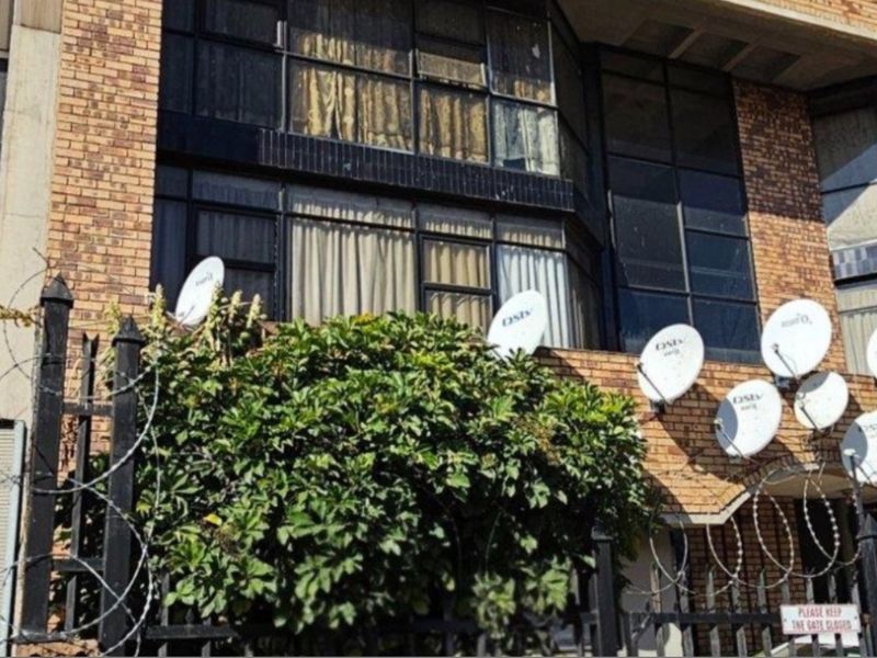 Front facade of a commercial brick building featuring large windows, security fencing with razor wire, and multiple satellite dish installations.