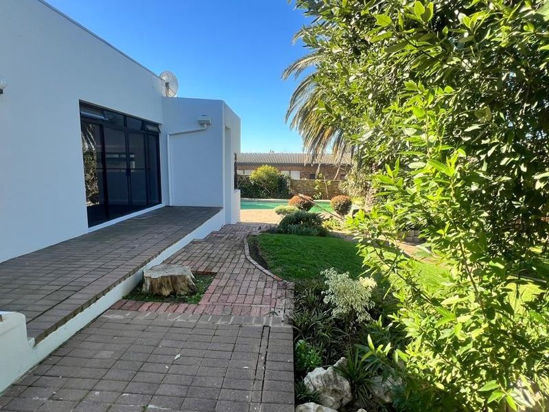 Rear exterior view featuring a paved patio area, brick pathways, landscaped garden, and a swimming pool visible in the background.