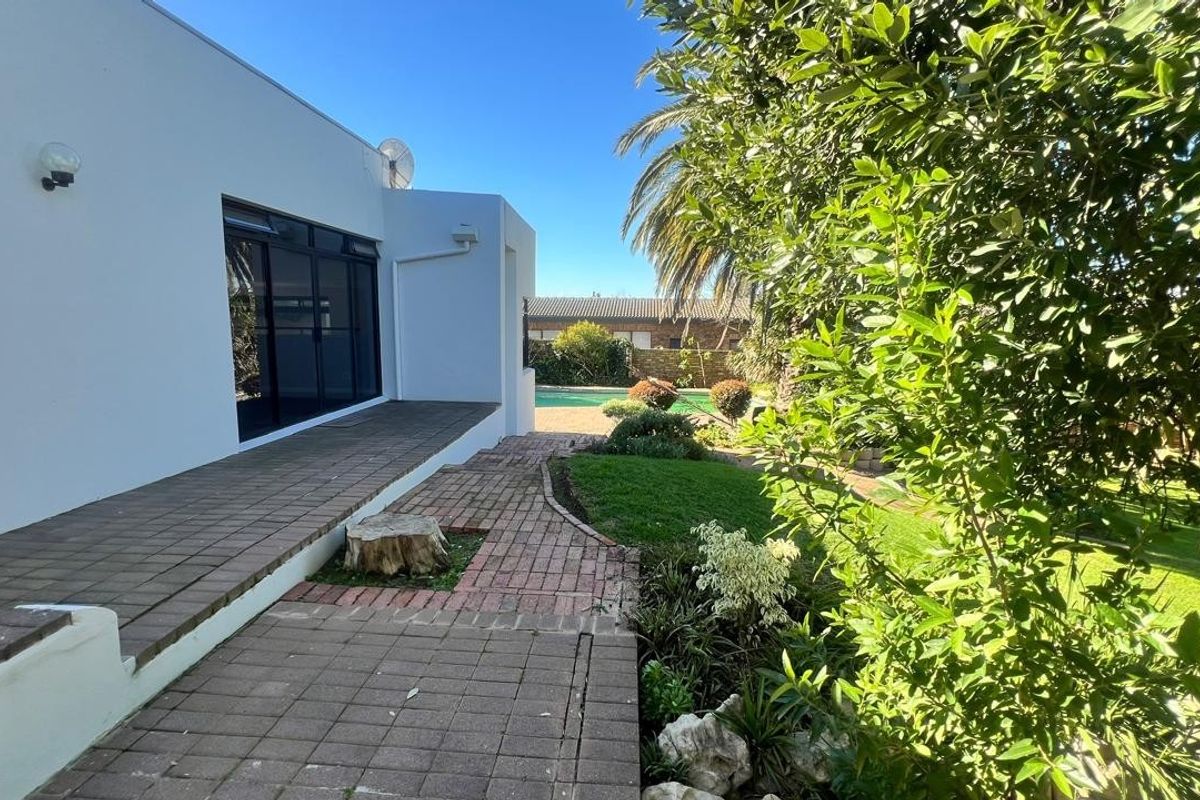 Rear exterior view featuring a paved patio area, brick pathways, landscaped garden, and a swimming pool visible in the background.