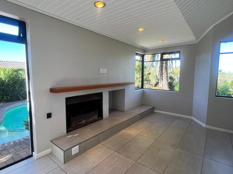A bright, empty living room featuring a built-in fireplace with a wooden mantel, tiled flooring, and large windows overlooking the swimming pool area.