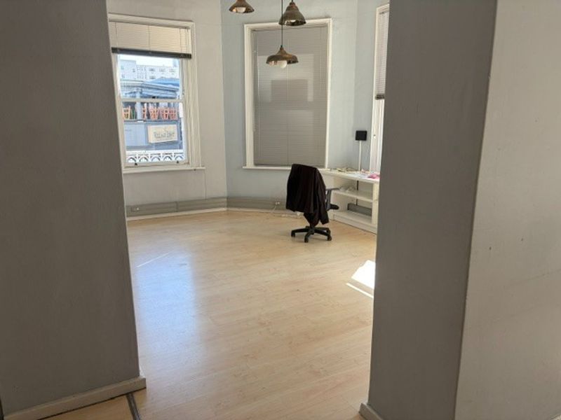 A bright, open-plan office space featuring light laminate flooring, large windows with venetian blinds, and built-in shelving units.