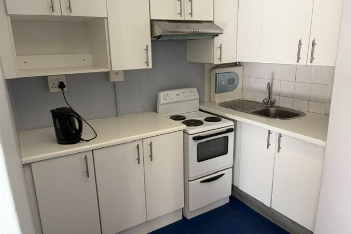Compact office kitchen featuring white cabinetry, a stainless steel sink, a freestanding stove with an extractor fan, and a tiled backsplash.