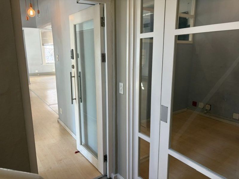 A bright hallway featuring glass panel doors, laminate flooring, and modern pendant lighting, leading into an open office space.