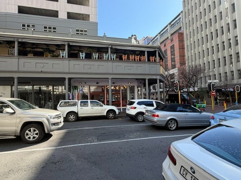Street-level view of a commercial office building featuring a prominent covered balcony and ground-floor storefront access in a busy urban setting.
