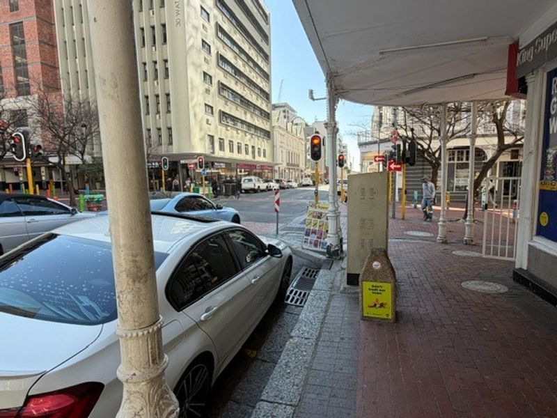 Street-level view of the office building exterior, featuring a covered pedestrian walkway, paved sidewalk, and urban infrastructure.