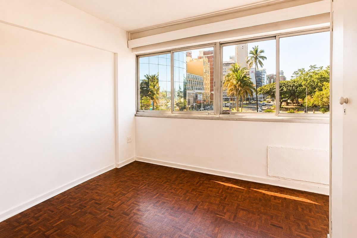 A bright, empty bedroom featuring classic parquet flooring, large windows with urban views, and a built-in cupboard.