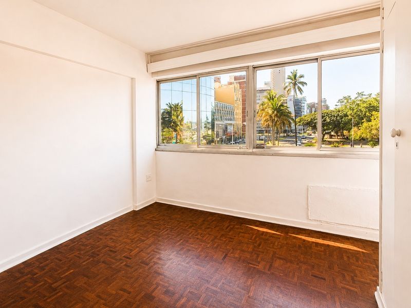 A bright, empty bedroom featuring classic parquet flooring, large windows with urban views, and a built-in cupboard.