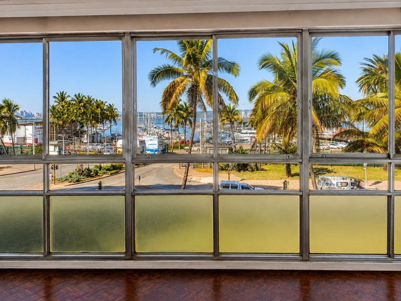 A bright interior space featuring expansive aluminium-framed windows offering a scenic view of the marina, palm trees, and coastal surroundings.