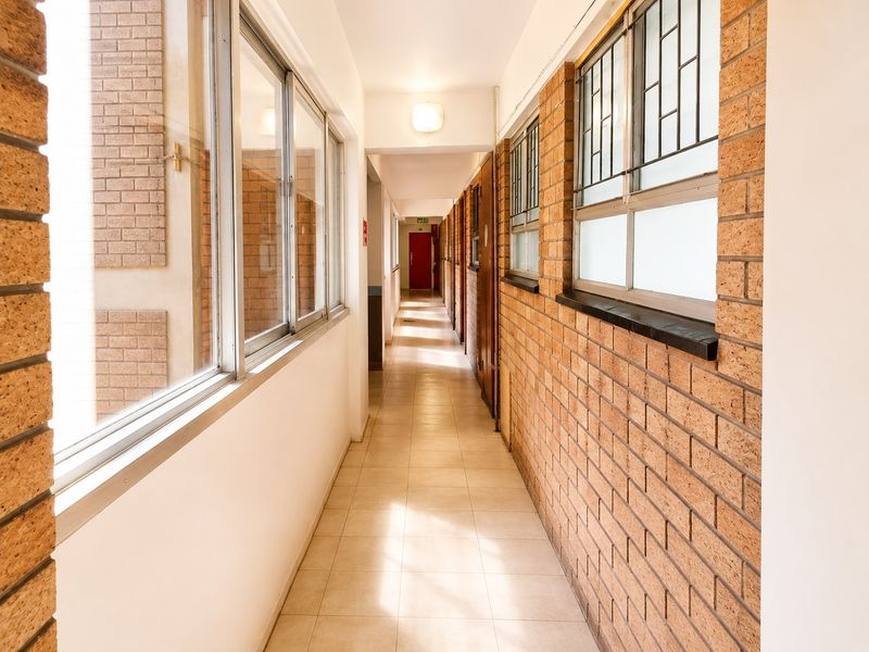 A bright, long apartment hallway featuring exposed brick walls, tiled flooring, and large windows with security bars.