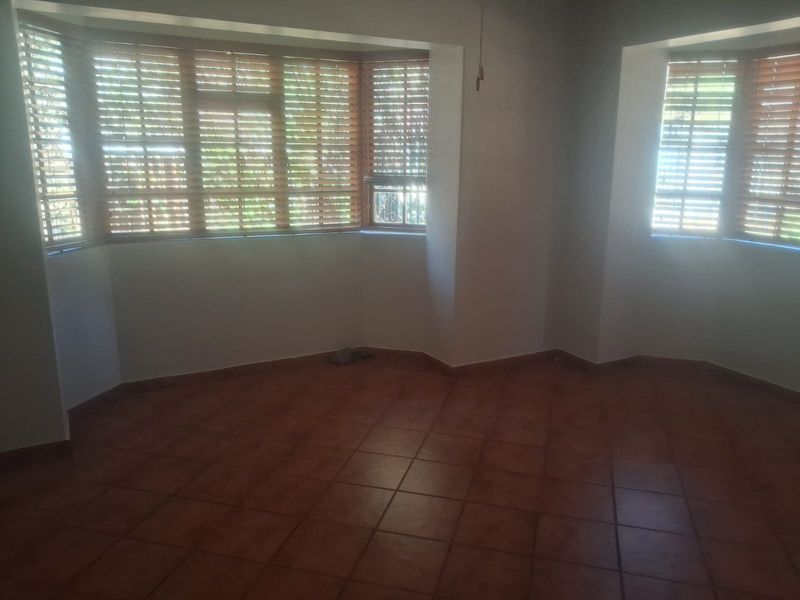 An empty living room featuring terracotta floor tiles and two bay windows fitted with wooden venetian blinds.