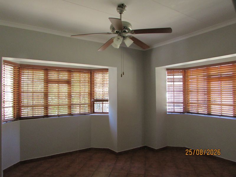 A bright, empty living room featuring tiled flooring, a ceiling fan, and dual bay windows fitted with wooden venetian blinds.