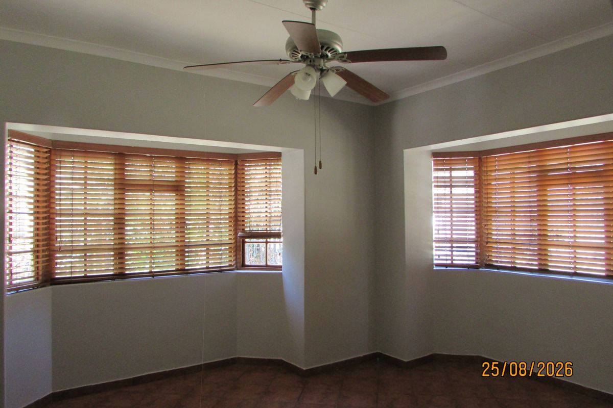 A bright, empty living room featuring tiled flooring, a ceiling fan, and dual bay windows fitted with wooden venetian blinds.
