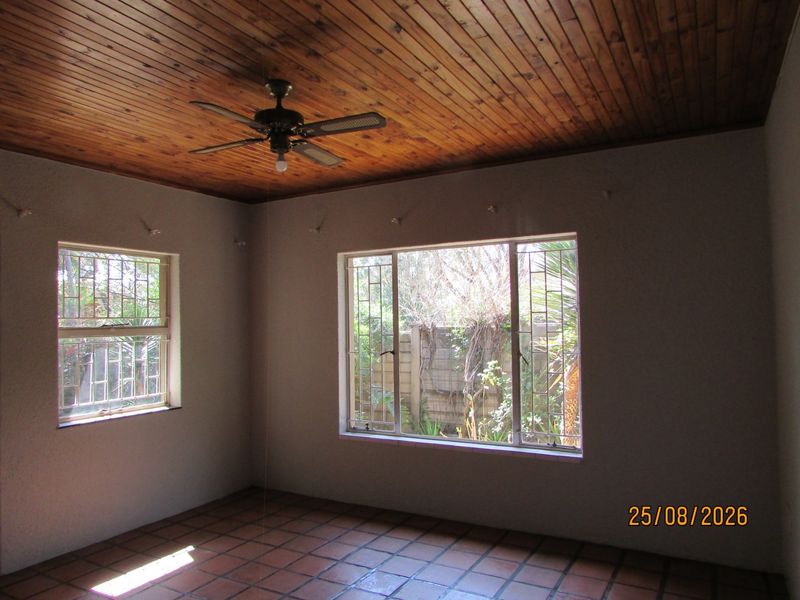 An empty living room featuring a warm timber-clad ceiling, tiled flooring, and large windows with security bars overlooking the garden.
