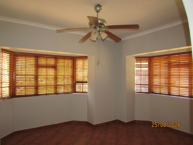An empty living room featuring tiled flooring, a ceiling fan, and large bay windows fitted with wooden venetian blinds.