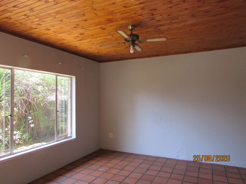 An empty living room featuring a timber ceiling, ceiling fan, and terracotta floor tiles, with large windows fitted with security bars.